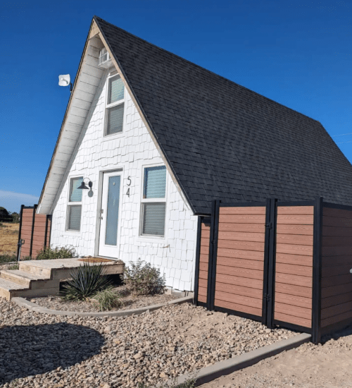 A white A-frame house with a black roof, surrounded by a gravel yard and wooden fencing, under a clear blue sky.